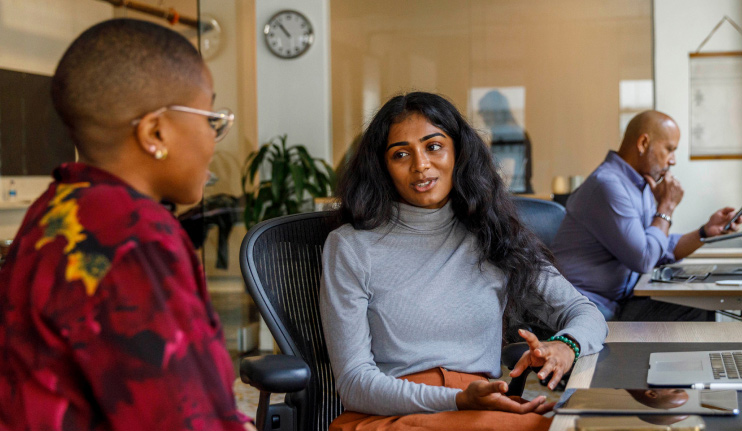 Two women talk together while sitting at a desk in their workplace