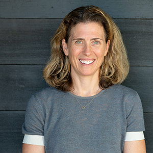 Portrait of a woman with shoulder-length blond hair wearing a grey shirt smiling in front of a grey wood wall.