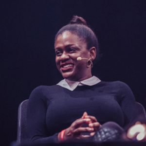 A Black woman wearing a black sweater with a white collared shirt sits on a stage wearing a microphone in front of a black background.