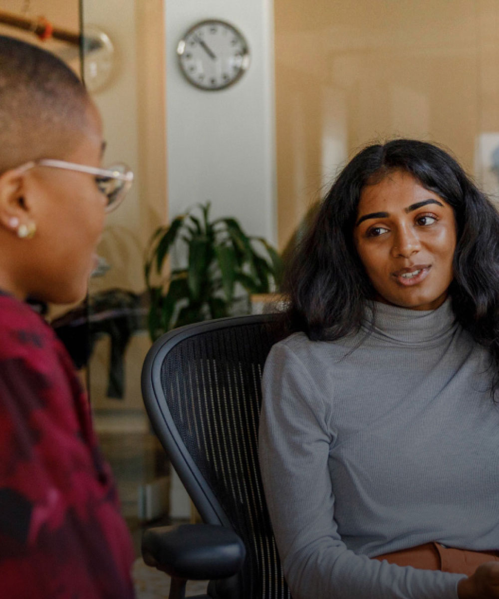 Two women talk together while sitting at a desk in their workplace.