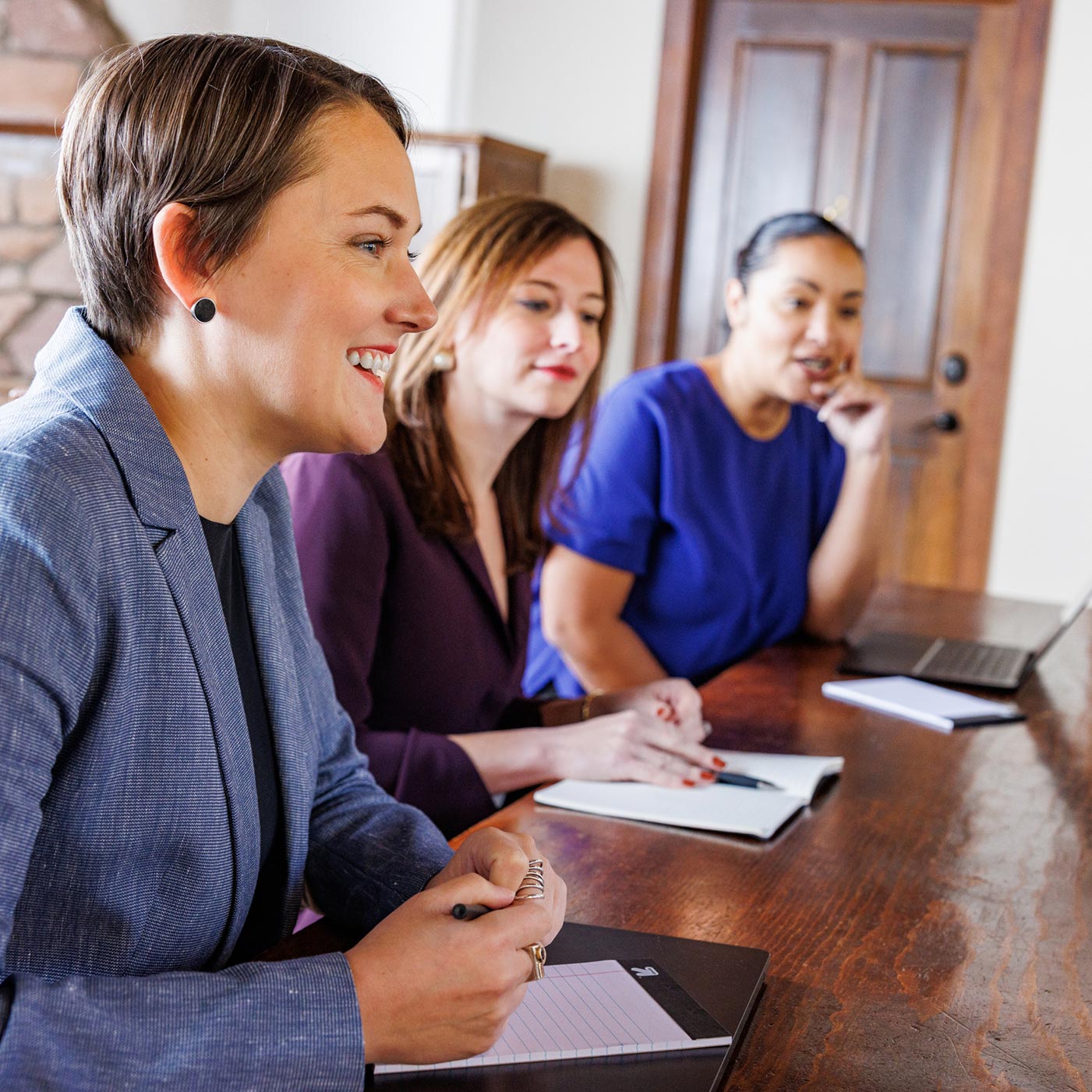 Three women working together at a table in the office, talking to a coworker out of frame.