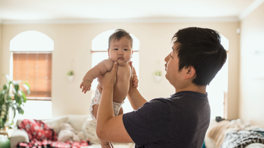 A father stands in his living room, holding his young baby up in the air