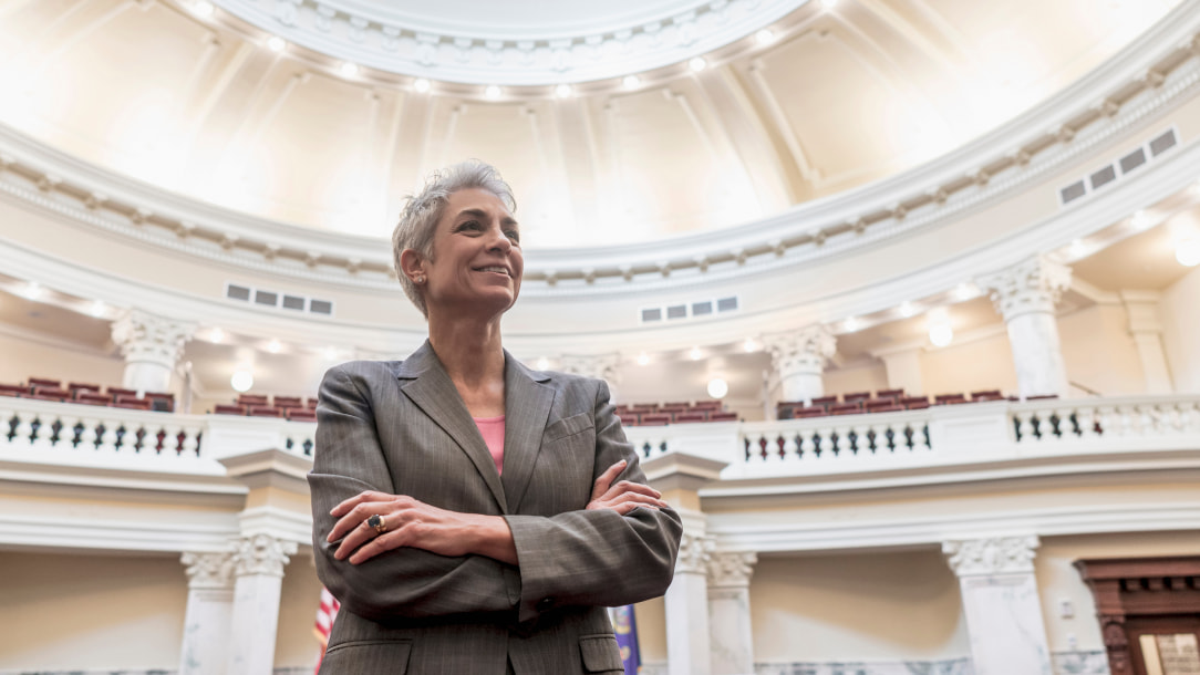 A female politician stands in a capitol building with her arms crossed.