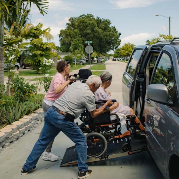 A man and woman work together to push a woman in a wheelchair up a ramp and into a vehicle.