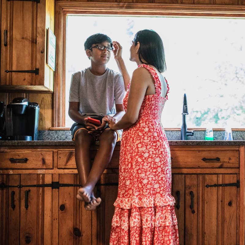A boy sits on a kitchen counter while his mom brushes his hair aside.