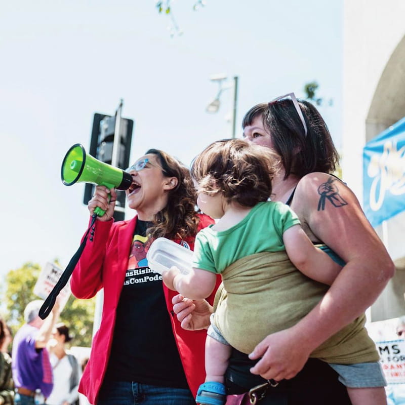 A woman speaks into a bullhorn at a rally next to a mom holding her young child.