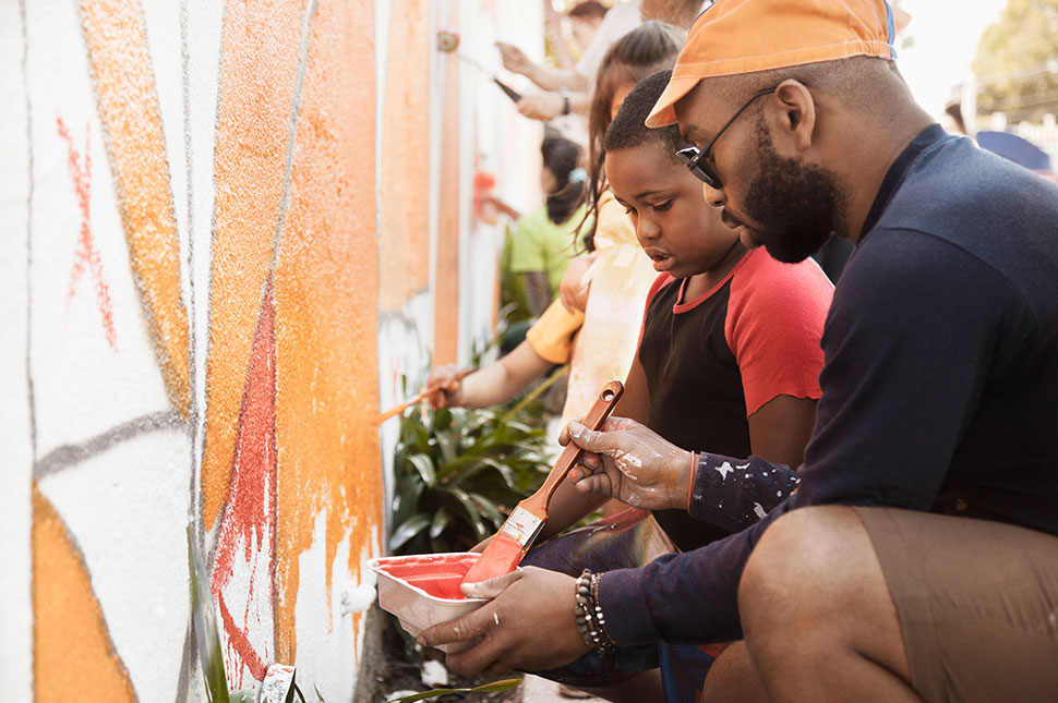 A black man helps a young black boy paint a mural on a wall at a community center