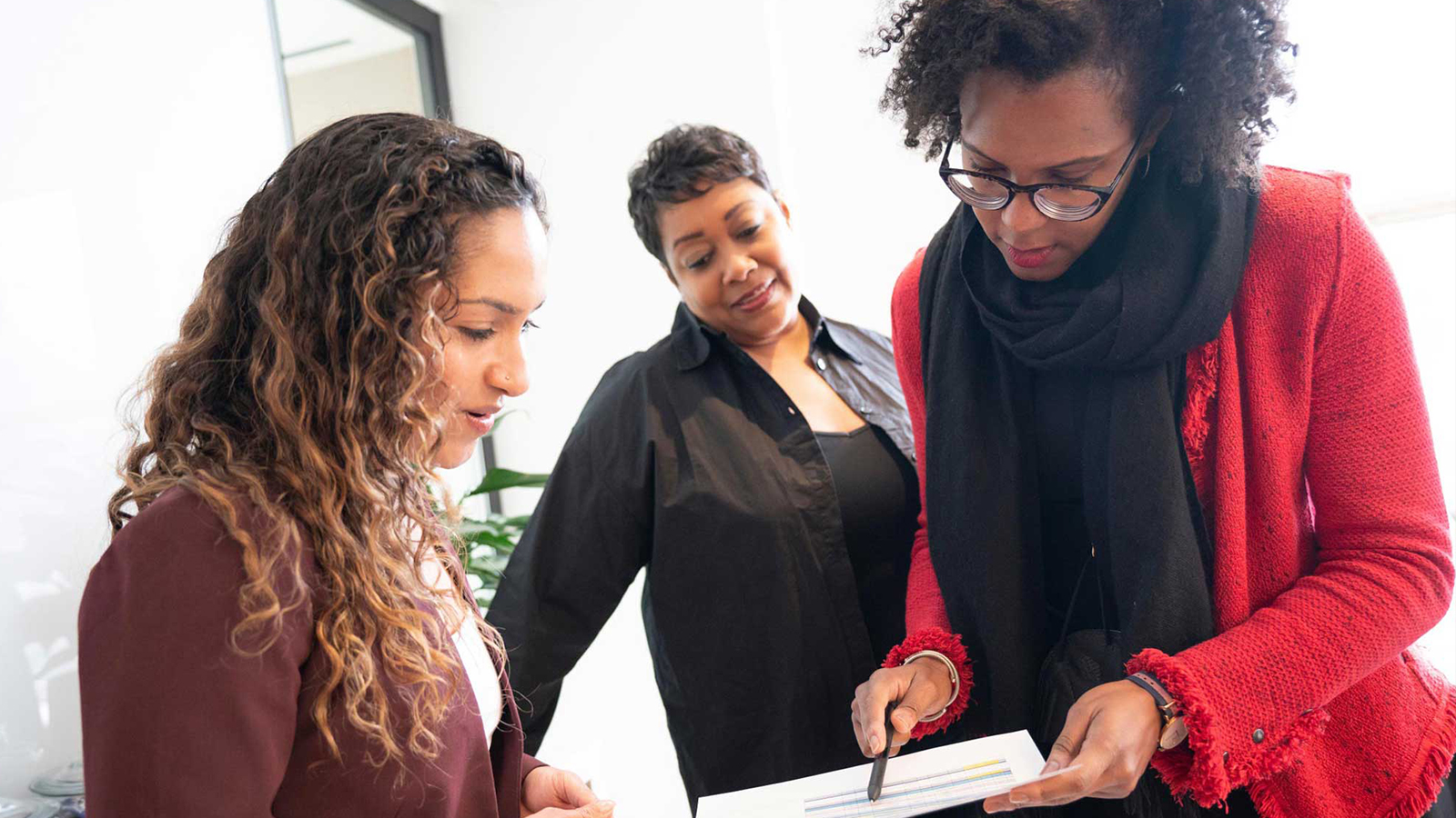 A woman wearing a red sweater and black scarf reviews a document with two colleagues.