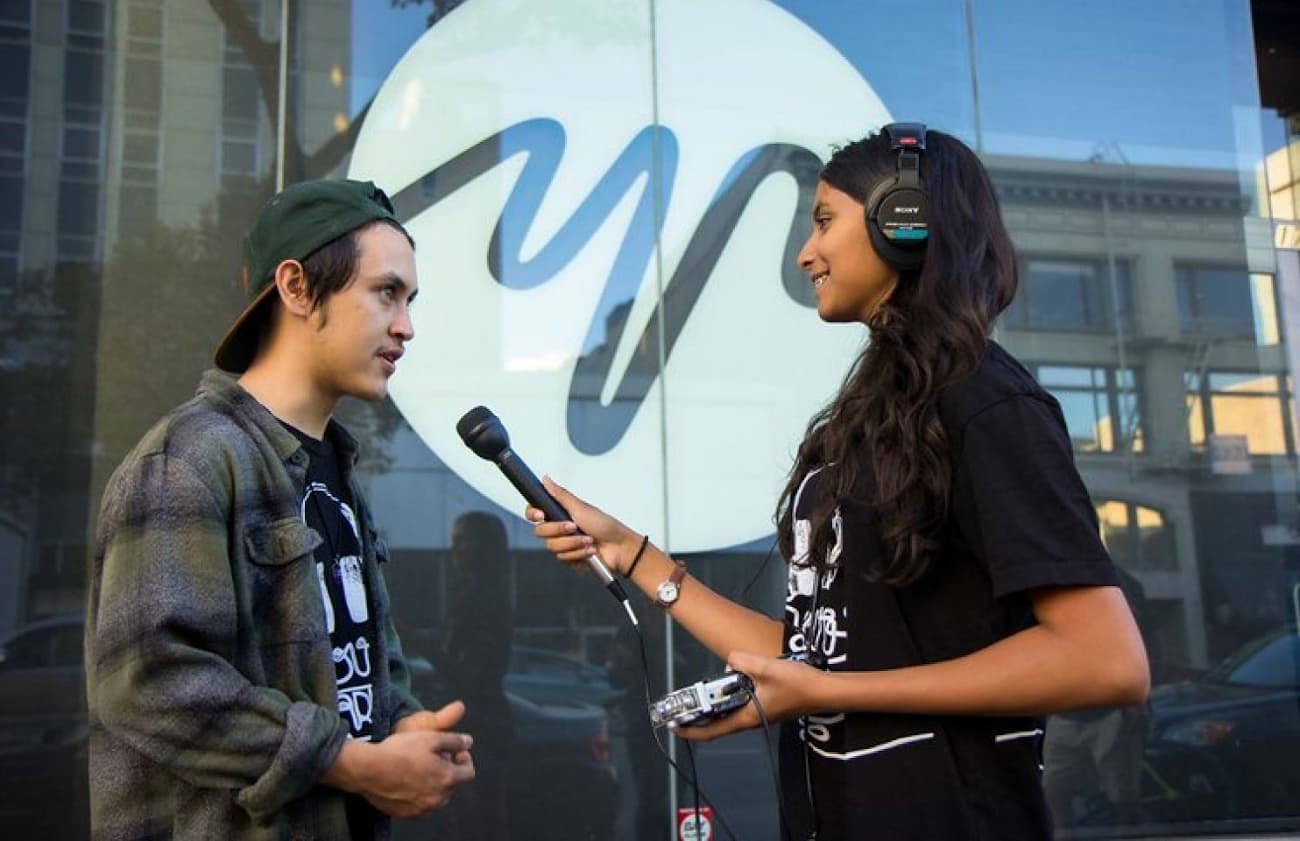 A young woman with headphones and a microphone interviews a young man for Pivotal Ventures partner YR Media.