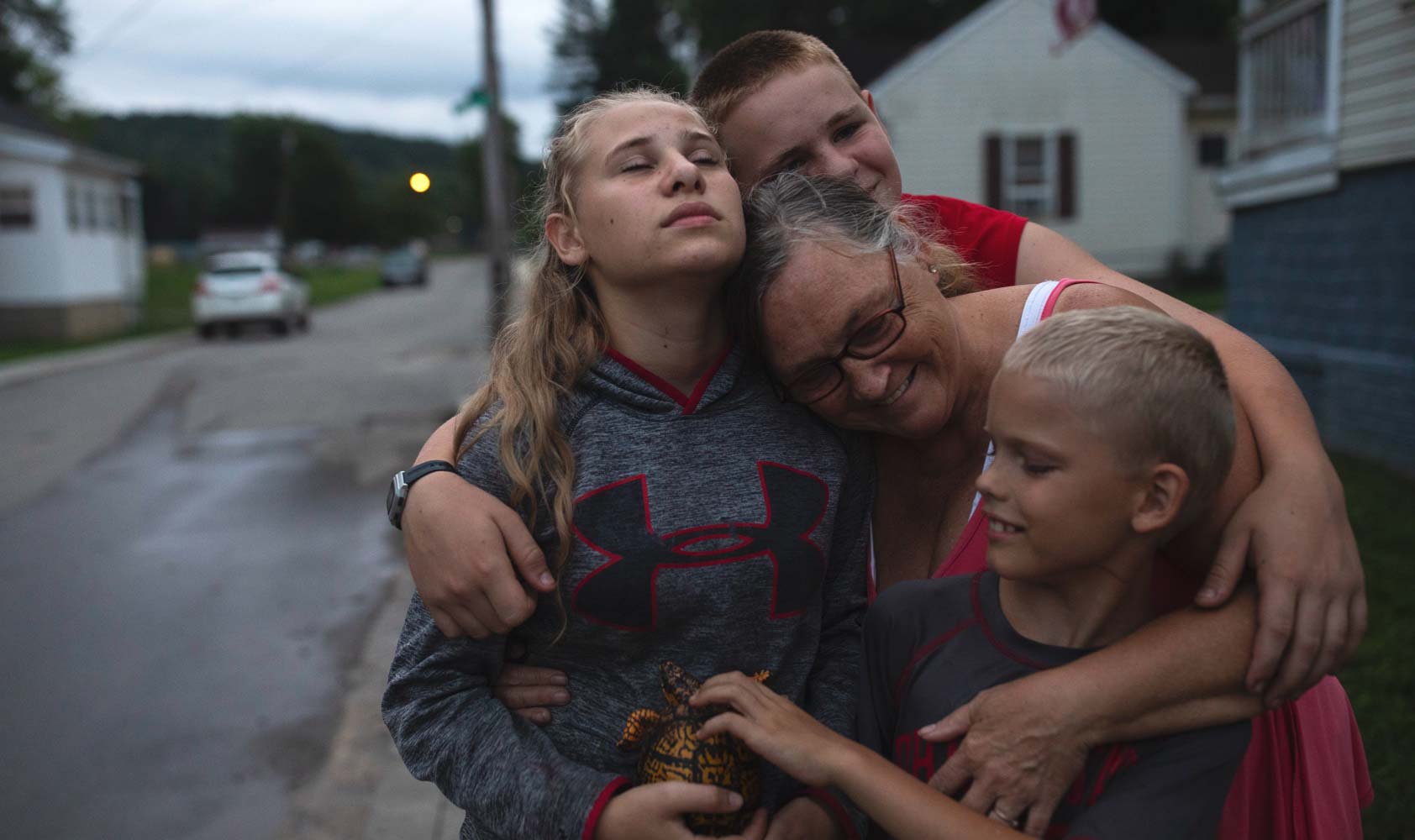 A 59-year-old woman and her three grandchildren, ages 9-14, stand on the street and embrace.