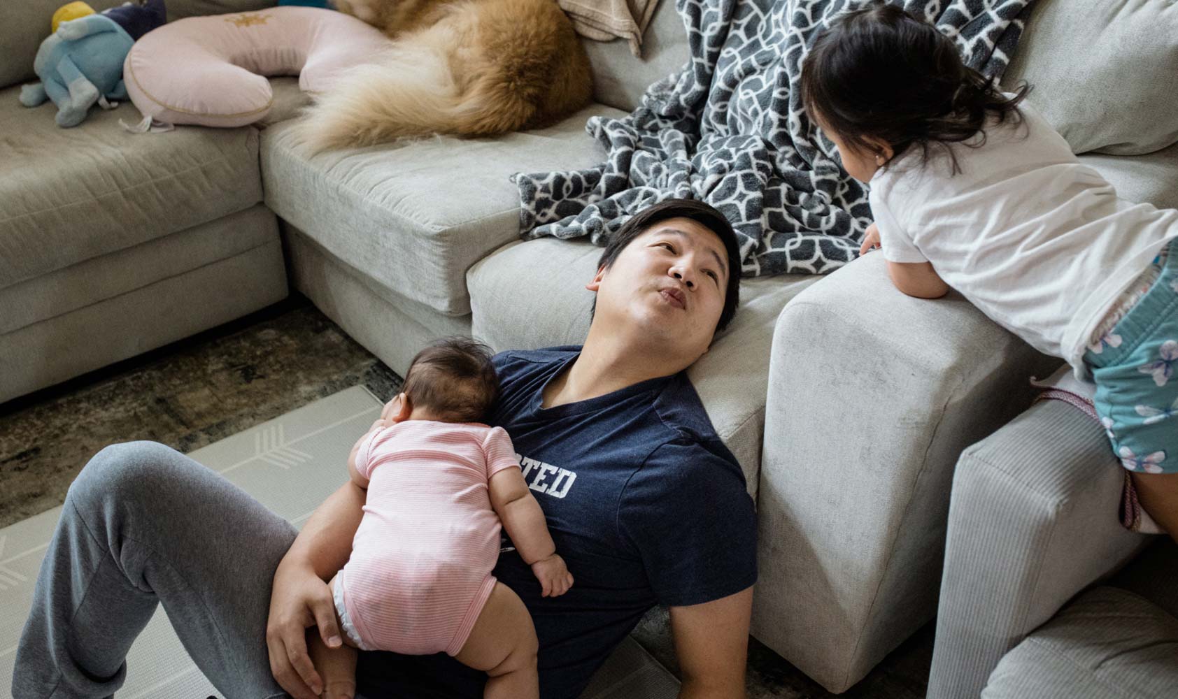 A 35-year-old man holds his baby and talks to his toddler while sitting on the floor leaning against a couch.