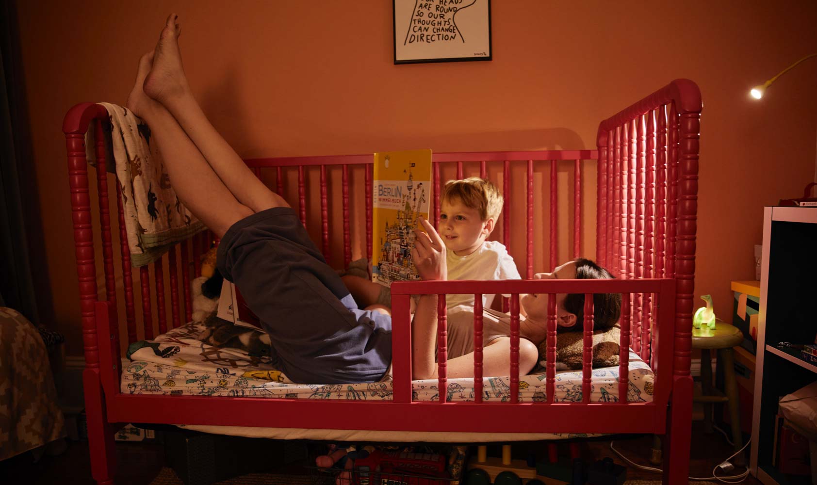 A 32-year-old woman lies in her 4-year-old son's small red bed reading him a book before bed.