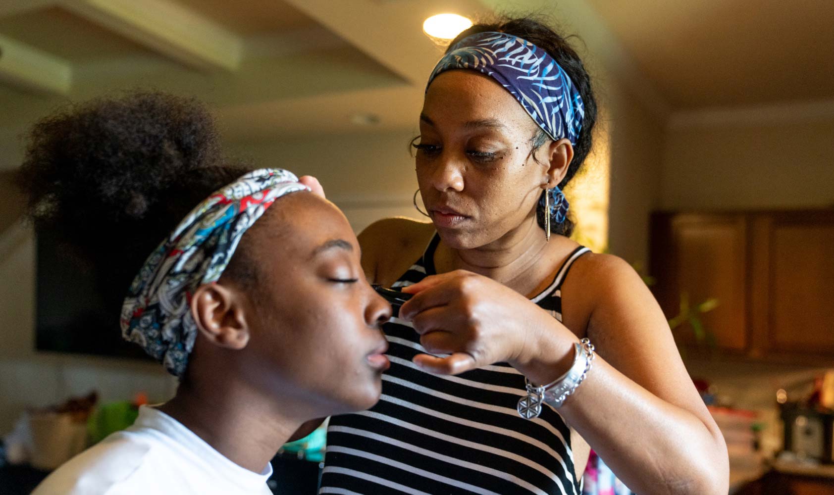 A 40-year-old woman stands in the kitchen helping her 17-year-old daughter, seated, to apply fake eyelashes.