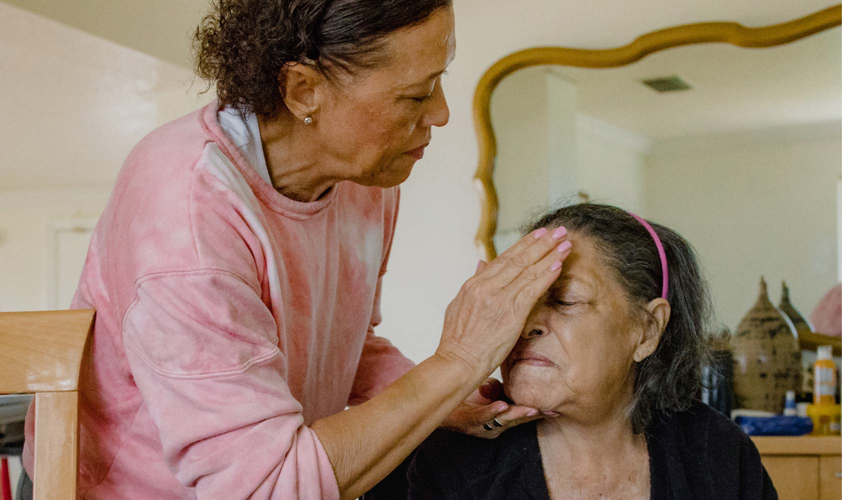 A 64-year-old woman leans over to apply moisturizer to her 97-year-old mother's forehead, with a mirror in the background.