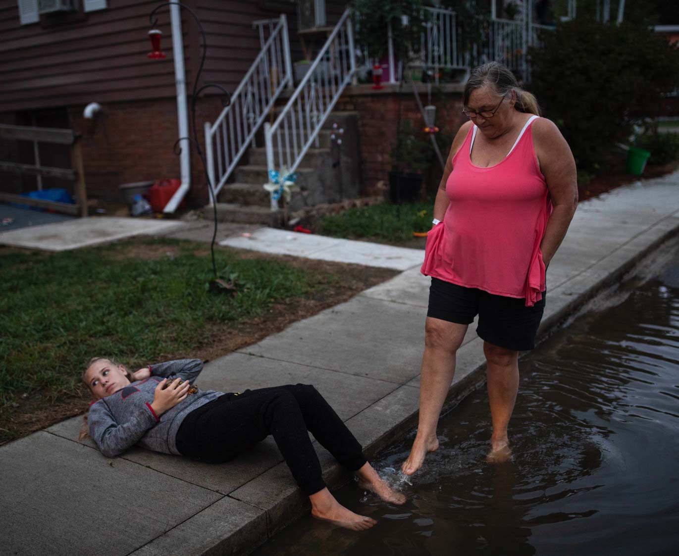 A 14-year-old girl lies on the sidewalk, holding a turtle, with her feet in a puddle. Beside her stands her 59-year-old grandmother, who uses her foot to playfully splash water on her granddaughter.