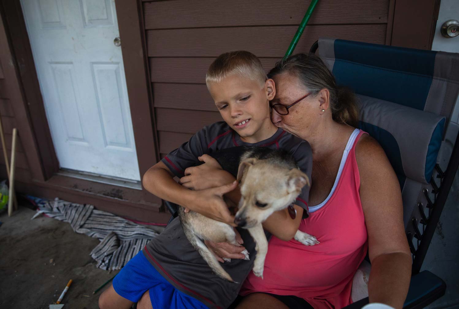 A 59-year-old woman sits in a chair on her porch with her young grandson and dog on her lap.