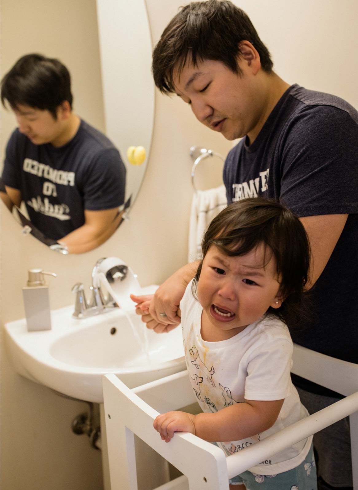 A 35-year-old father and his unhappy toddler stand together at the bathroom sink as he washes her hands.