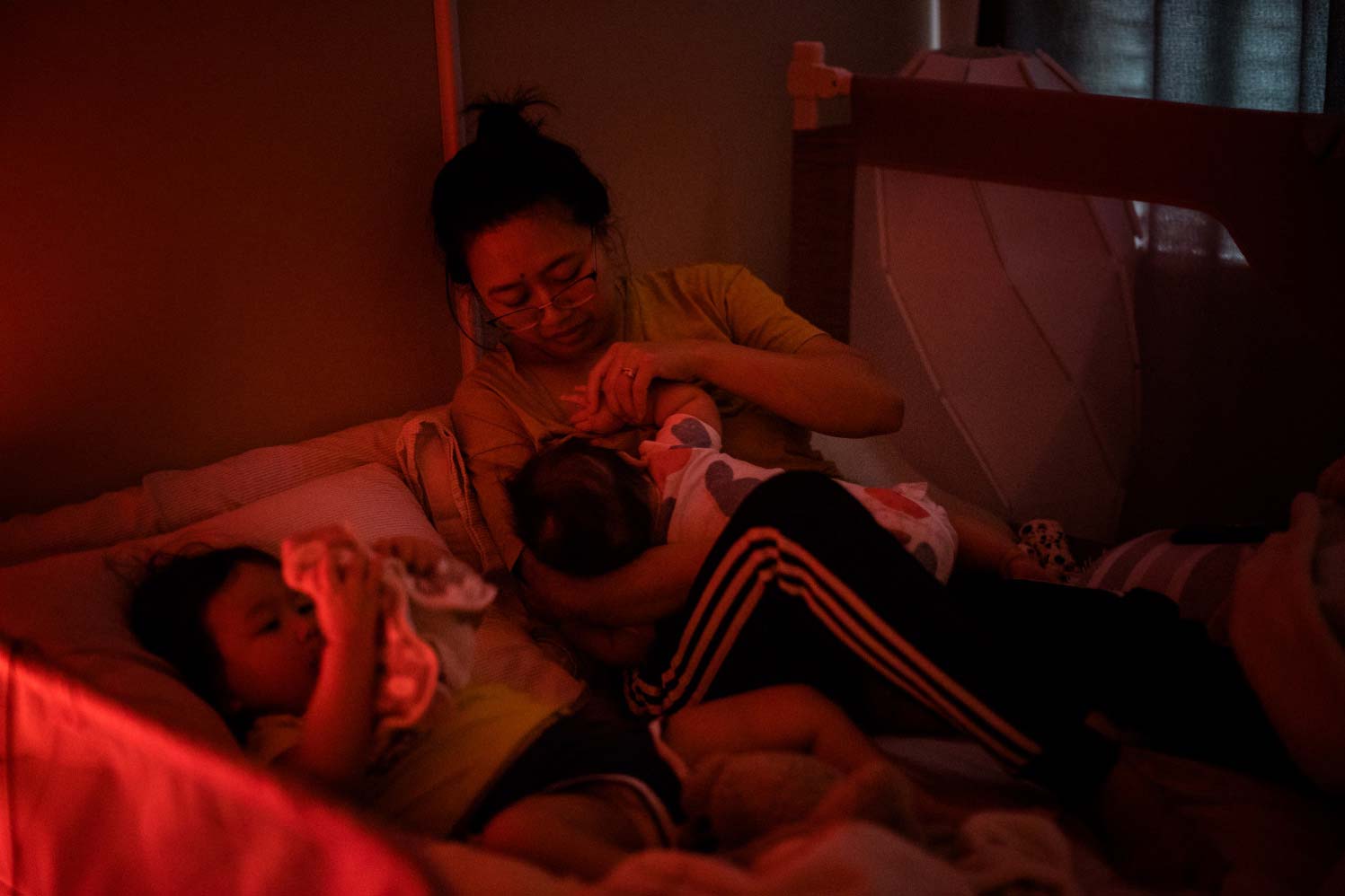 A 32-year-old mother lies next to her toddler in bed in a dark room while breastfeeding her baby.