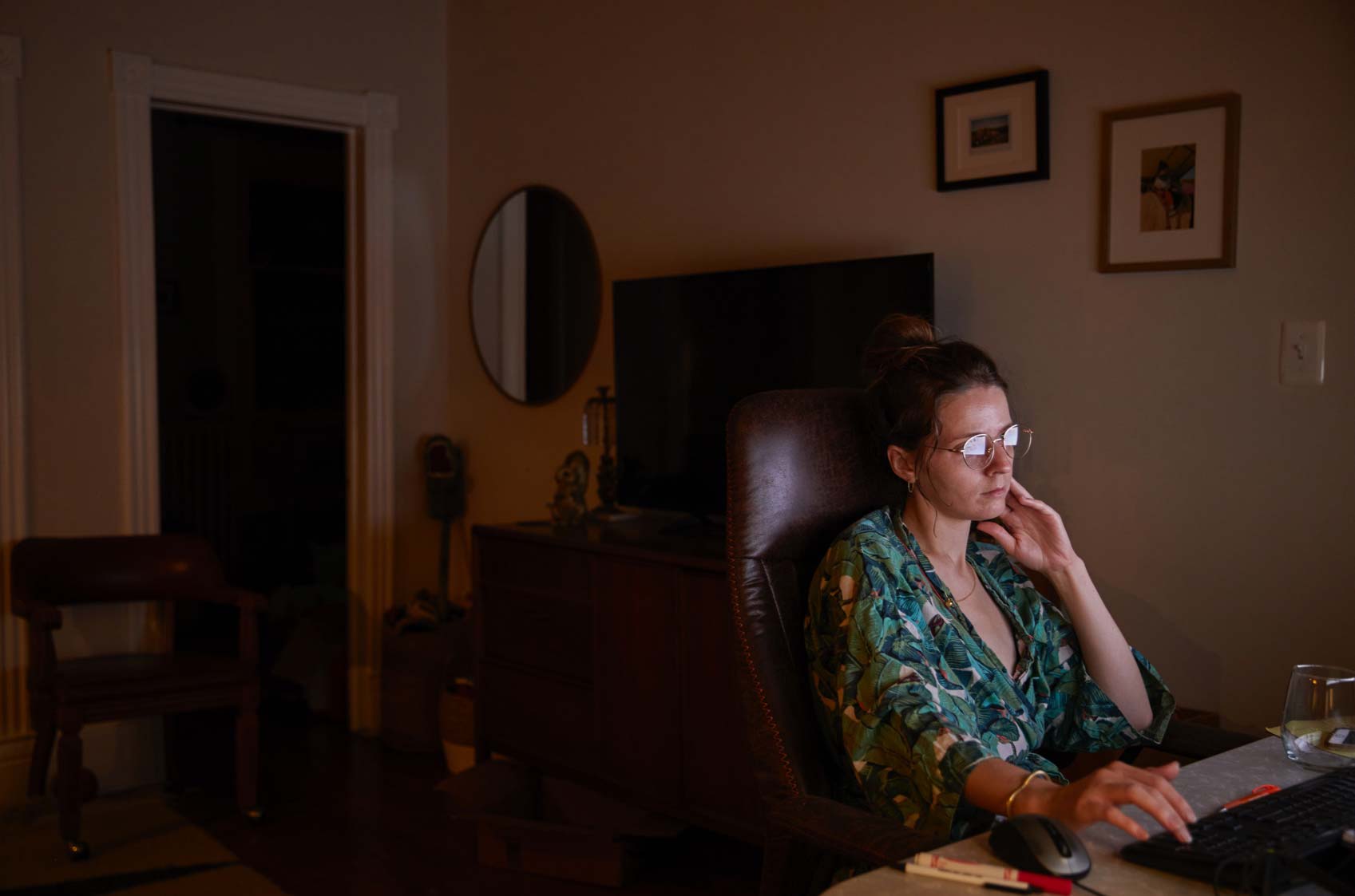 A 32-year-old woman sits at her desk in a dark room, her face lit up by the glow of her computer screen.