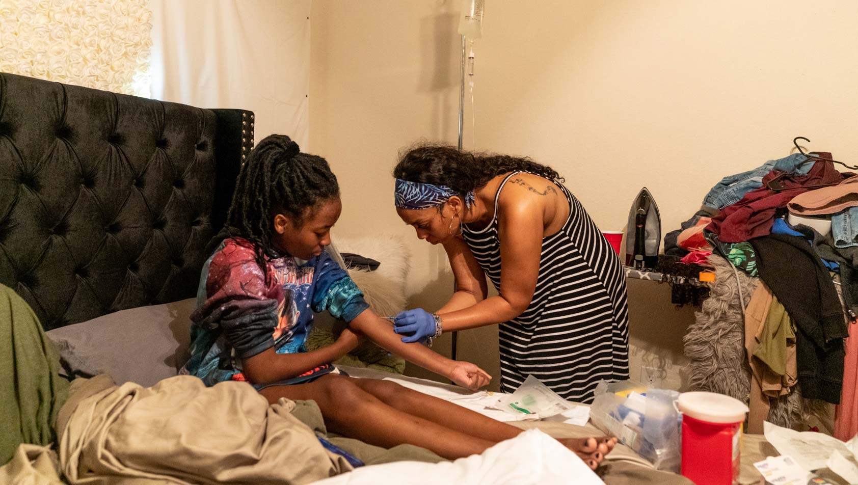 A 40-year-old woman bends down to give an IV to her 16-year-old daughter, who is sitting on a bed in their home.
