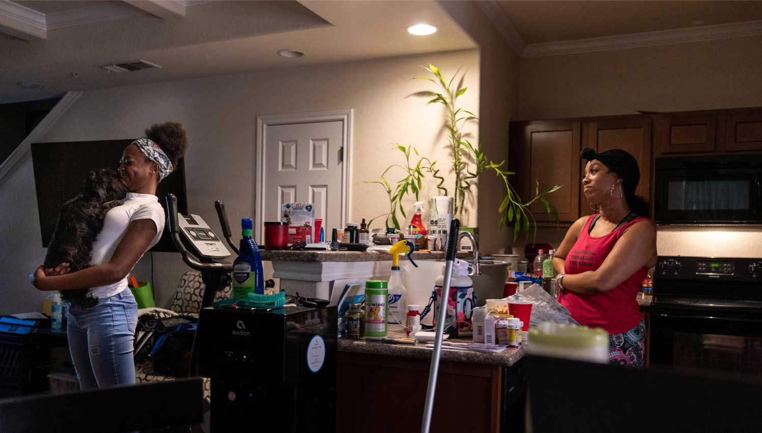 A 17-year-old girl holds a small dog in her arms as she stands in the kitchen talking to her 40-year-old mother, who is cooking.