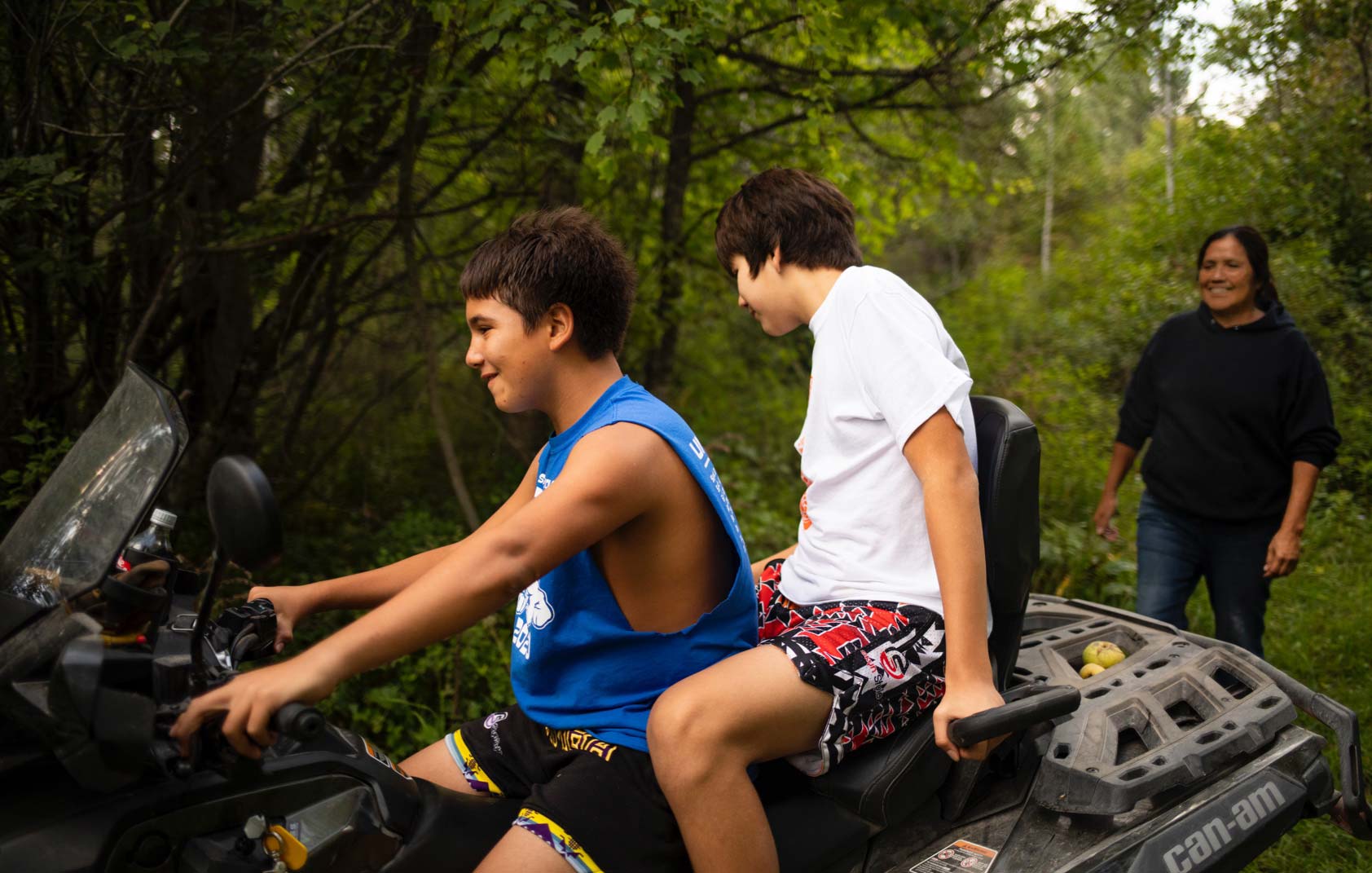 A 13-year-old boy drives a four-wheeler with another boy sitting behind him, as his 62-year-old grandaunt watches them, smiling.