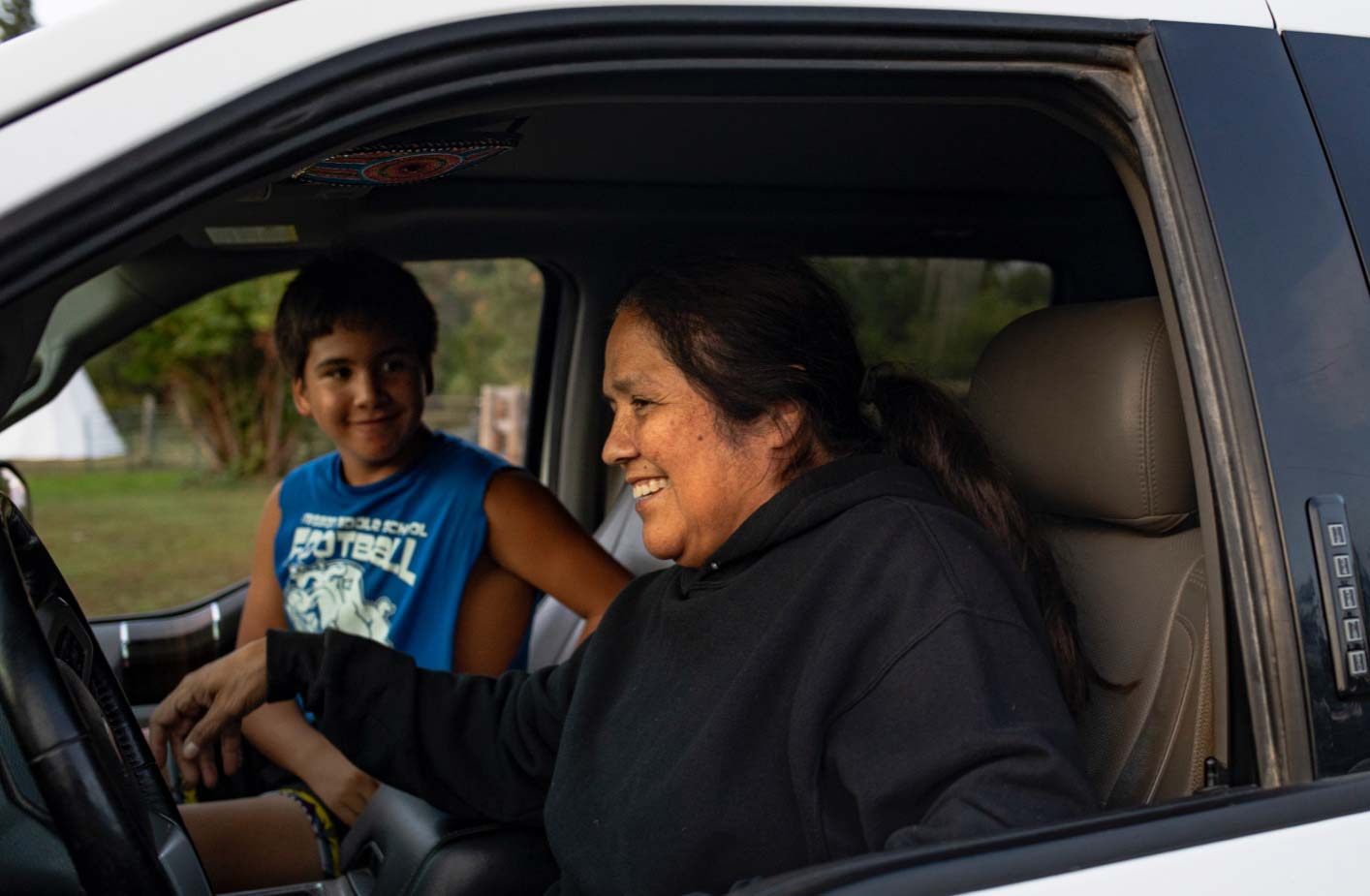 A 62-year-old woman sits in the driver's seat of a white vehicle beside her 13-year-old grandnephew; both are smiling.