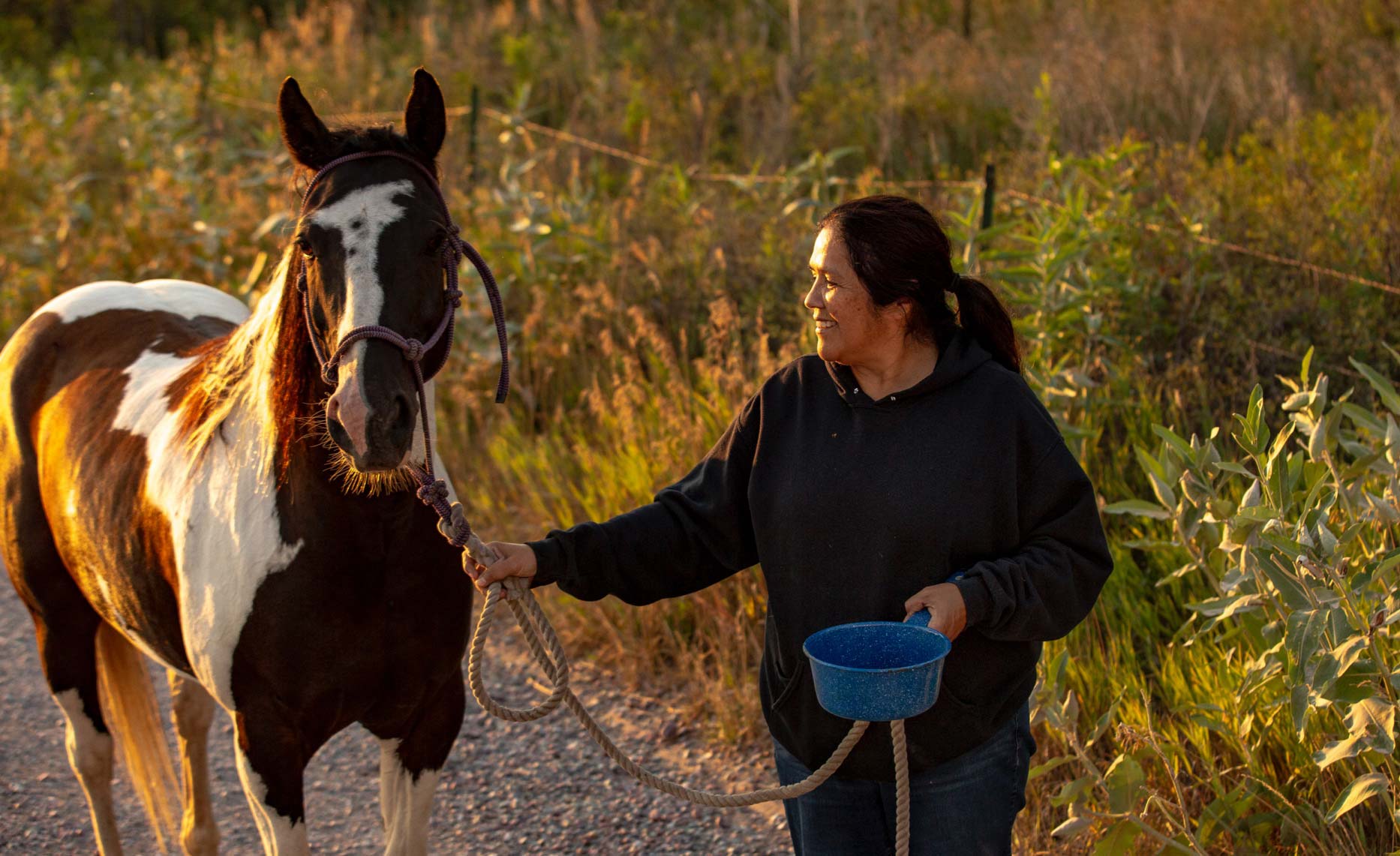 A 62-year-old woman walks beside a brown and white horse as she guides it by the reins, with a field of grass in the background.