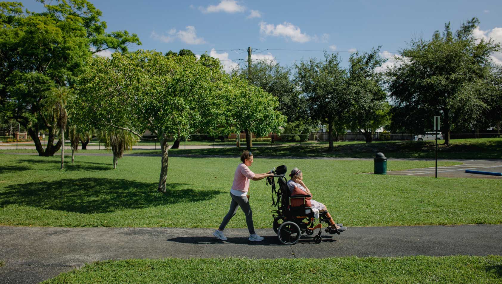 A 64-year-old woman pushes her 97-year-old mother's wheelchair on a paved path through a bright green park filled with trees.