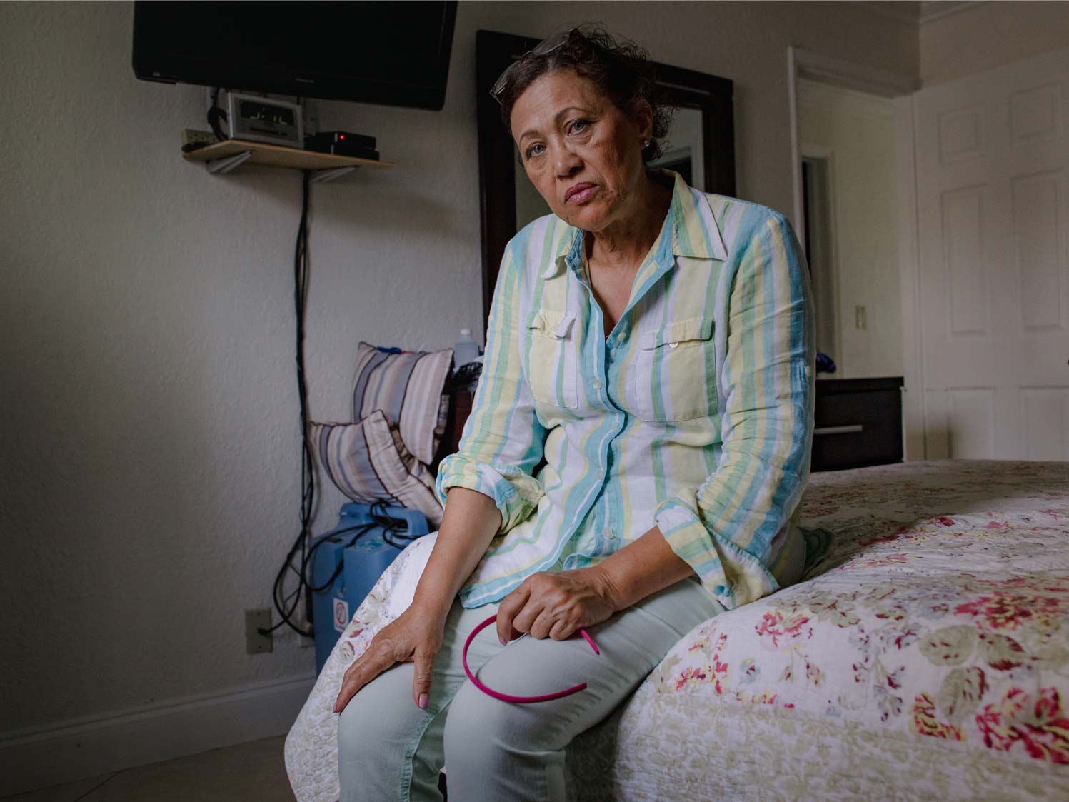 A tired 64-year-old woman sits on the edge of a bed covered in a floral bedspread.