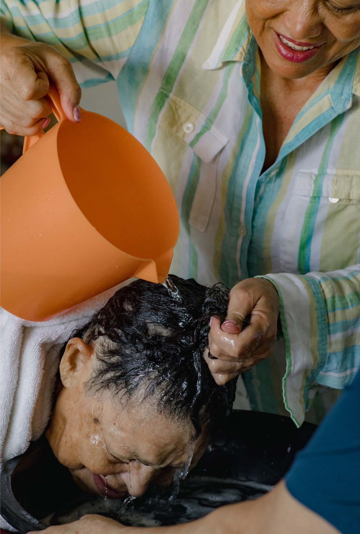 A 64-year-old woman pours water from an orange pitcher over the lowered head of her 97-year-old mother.