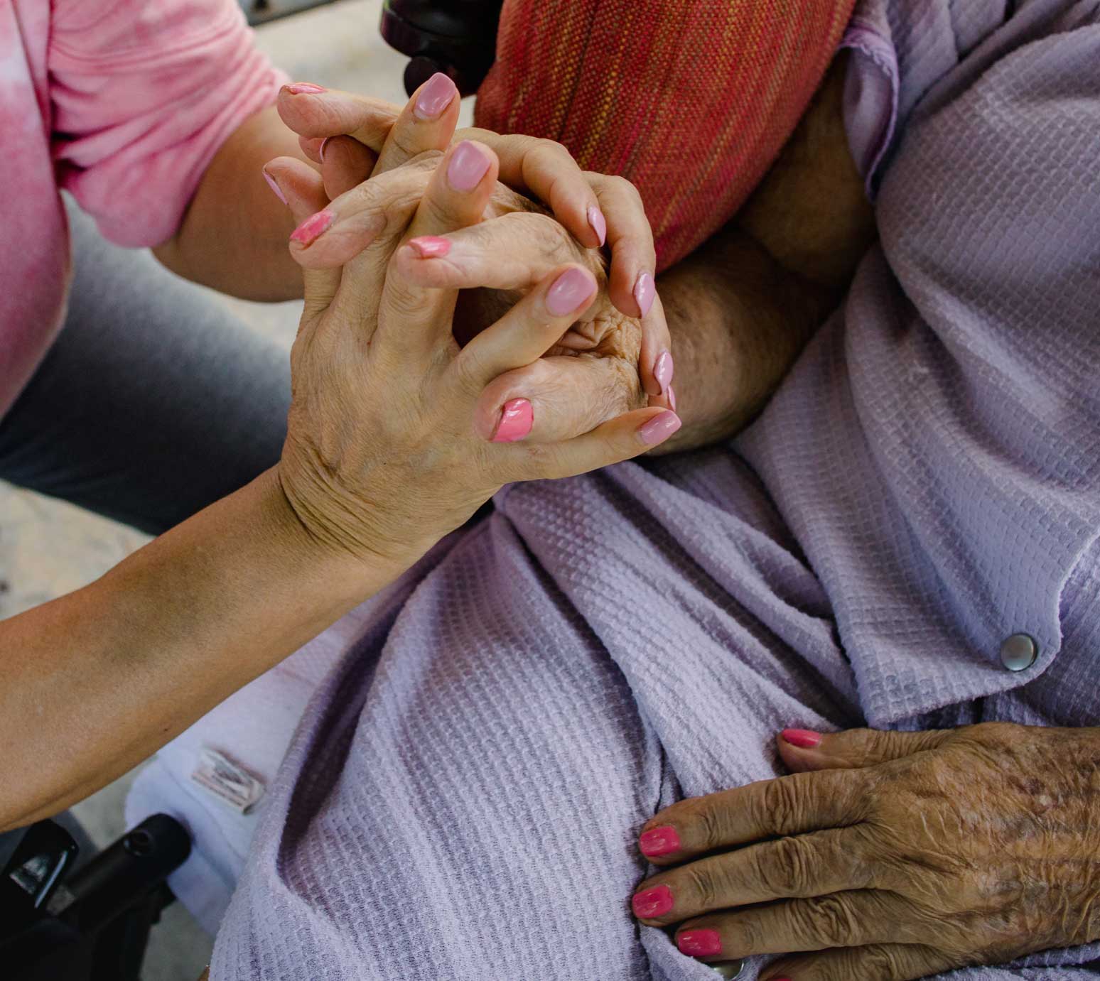 A 64-year-old woman's hand intertwines with that of a 97-year-old woman, whose other hand rests on her lap. The nails on both women's hands are painted.