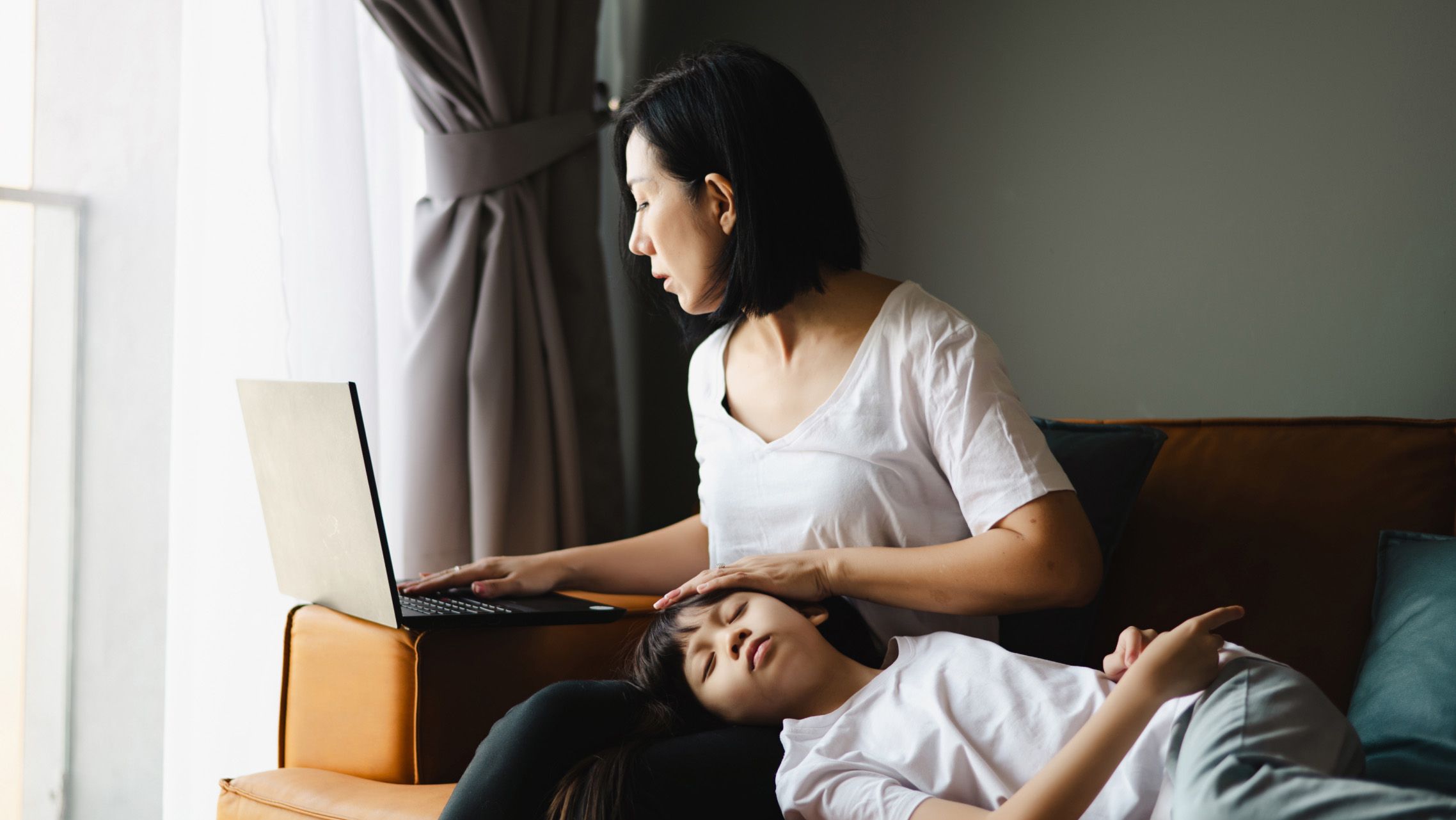 A mom tries to work on her laptop while her young son rests his head on her lap.