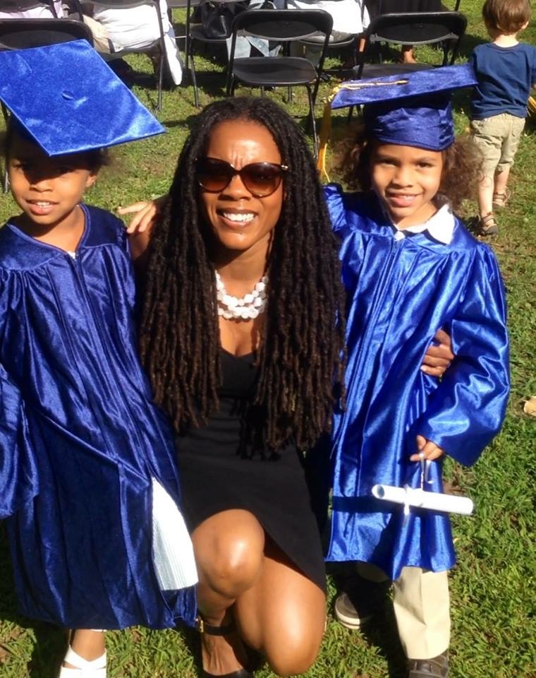 Dr. C. Nicole Mason wearing a black dress and sunglasses crouches next to her two young children who wear blue graduation gowns and caps.