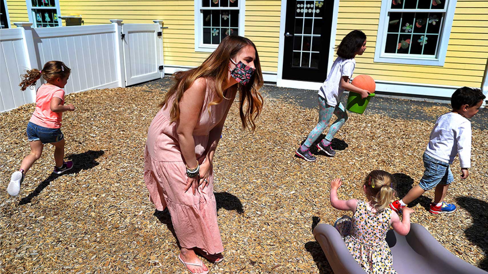 A daycare worker leans over and watches a child go down a slide with other children running around behind her.