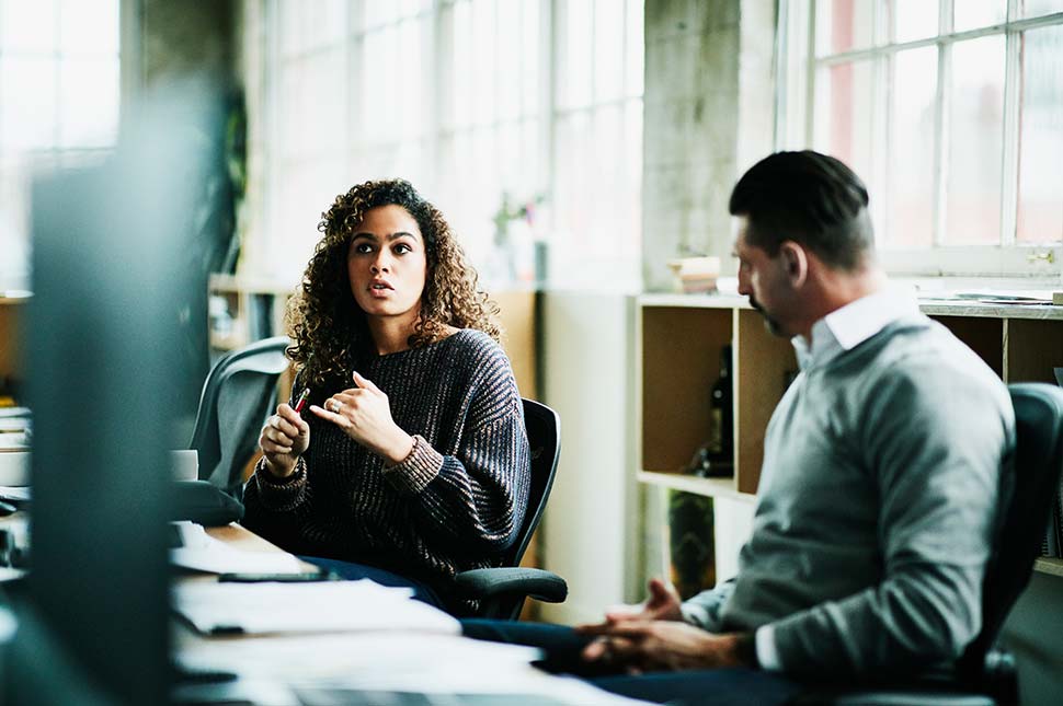 Two colleagues sit and discuss their work at a desk.