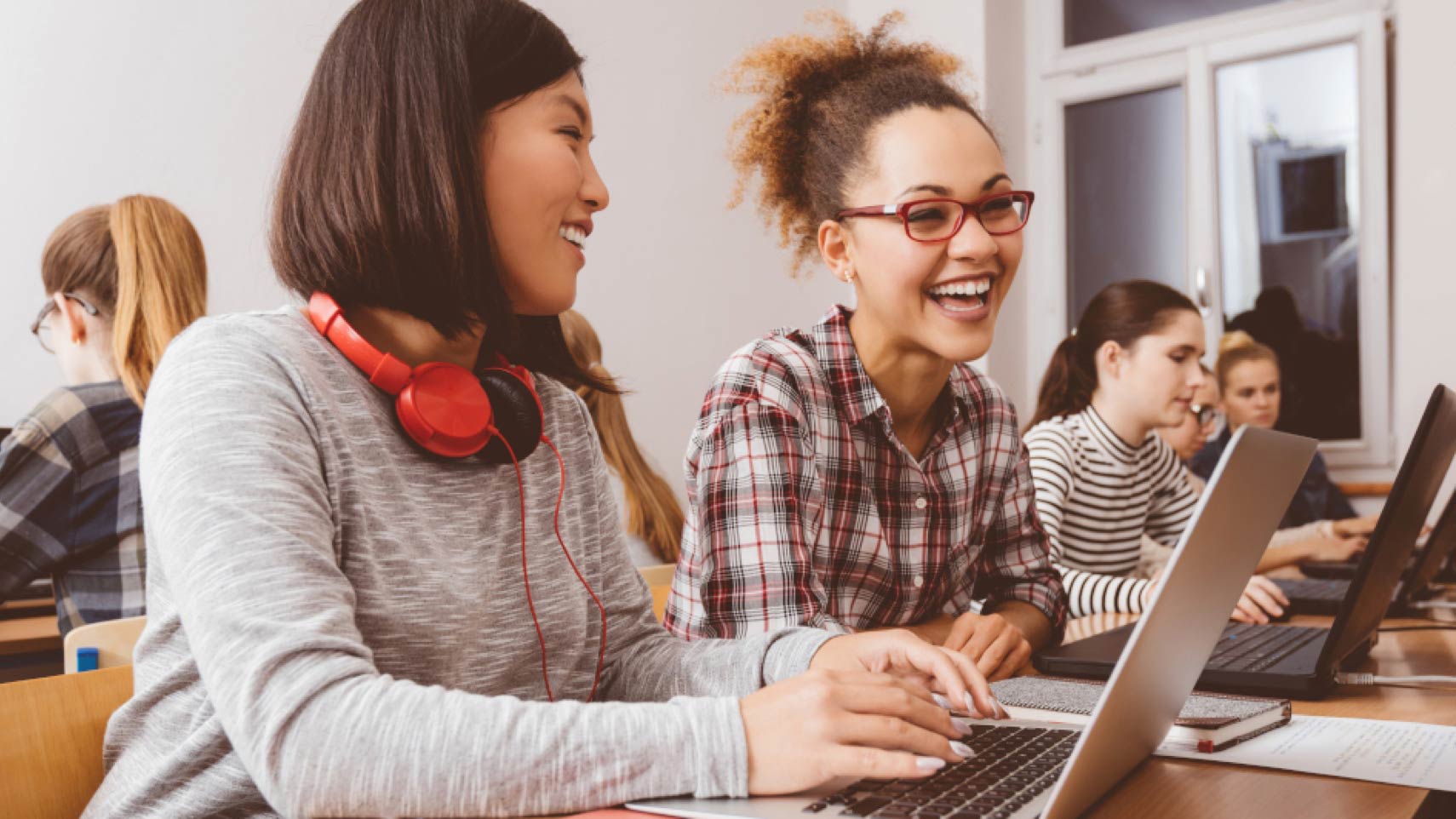 Two young people sit smiling in front of laptop computers, while others sit at computers nearby.