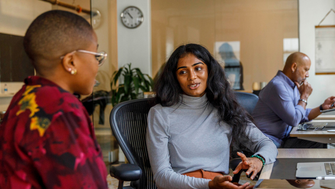 Two women talk together while sitting at a desk in their workplace.