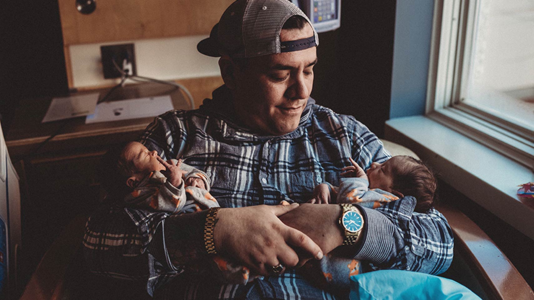 A person sits holding a baby in each arm and smiles down at one of them.