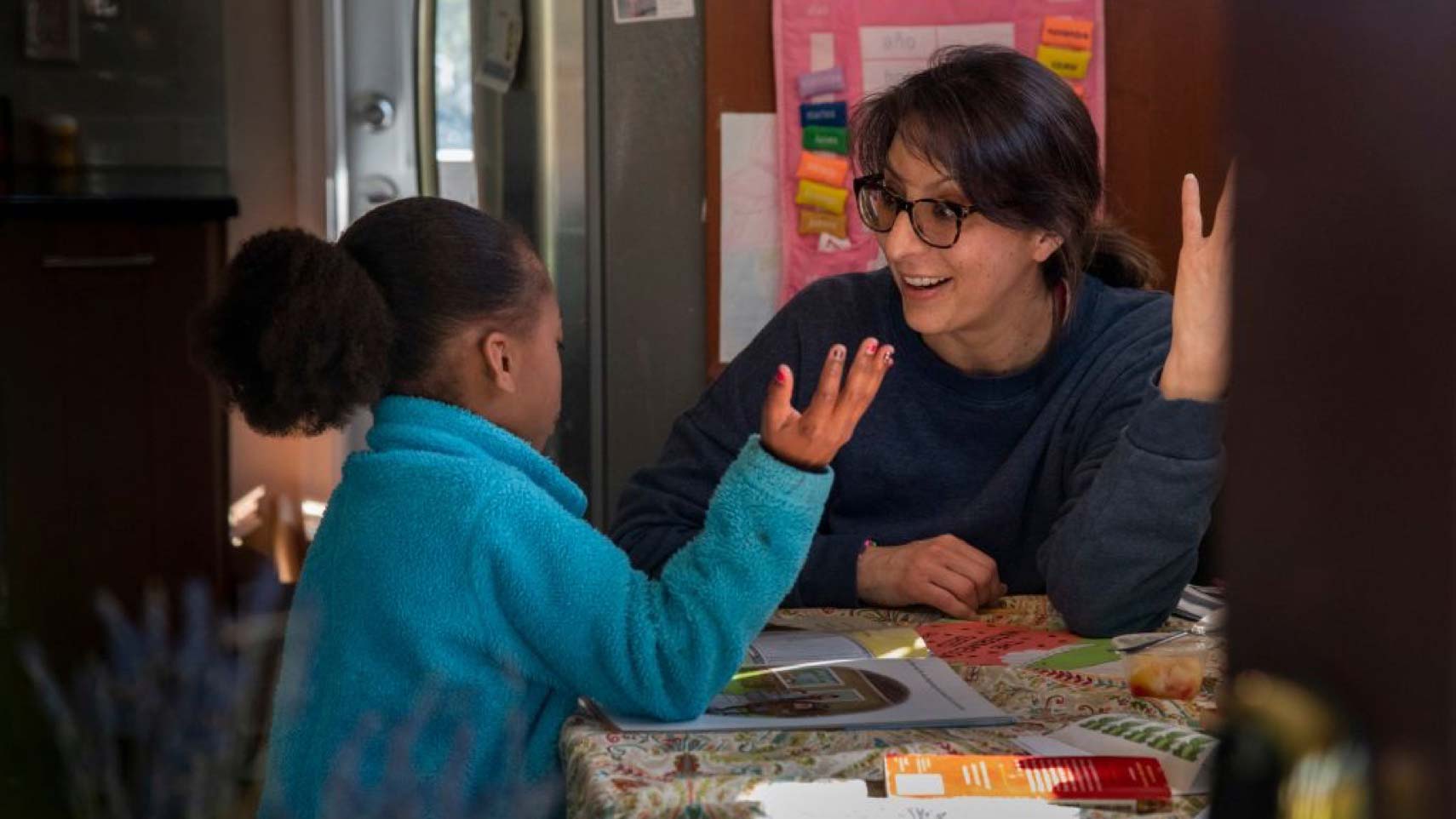 A woman with an animated expression sits at a table talking to a child. In the background, there is a pink poster that says "Mi Calendaro" in colorful letters.