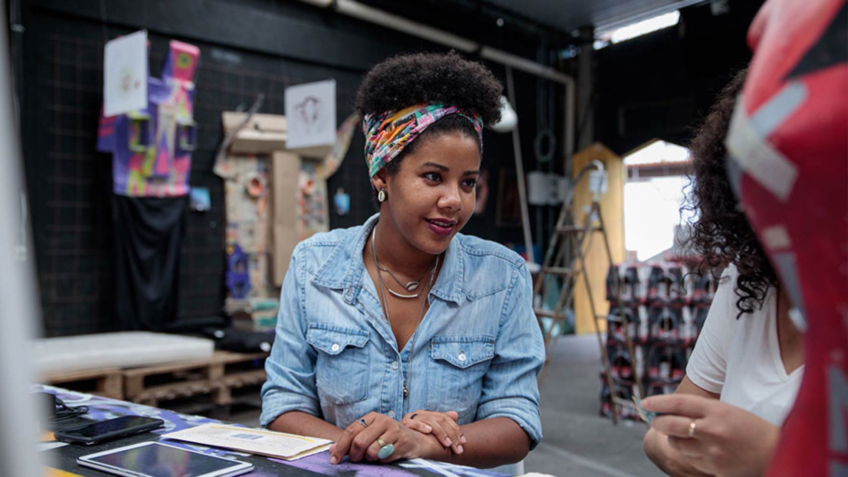 A person sits at a table talking to someone, with glimpses of artwork in the background.