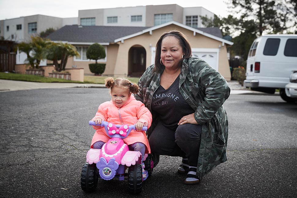 Anita crouches in her cul de sac with her daughter, who is riding a toy bike