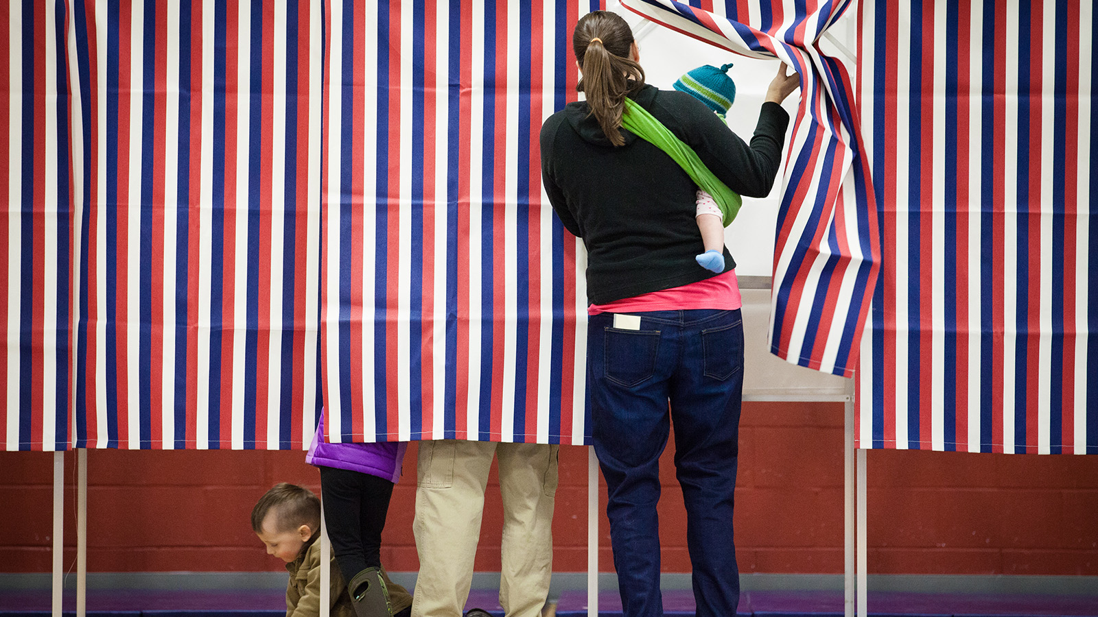 Woman with young child enters voting booth through red, blue, and white curtain.
