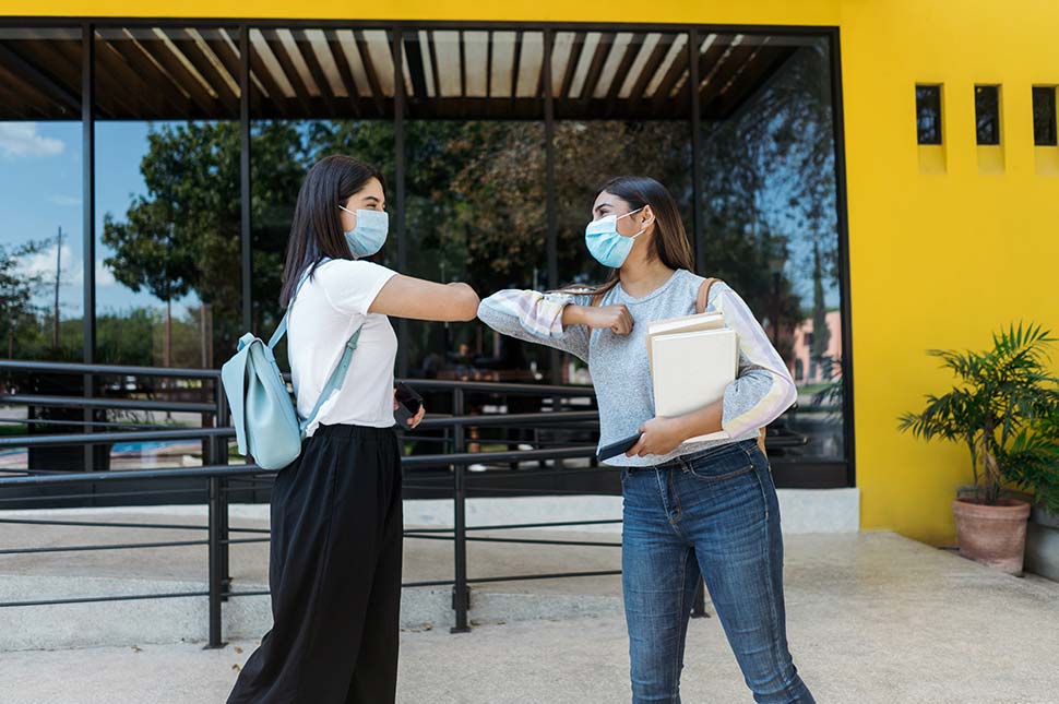 Two women wearing basks and backpacks bump elbows outside a yellow building.