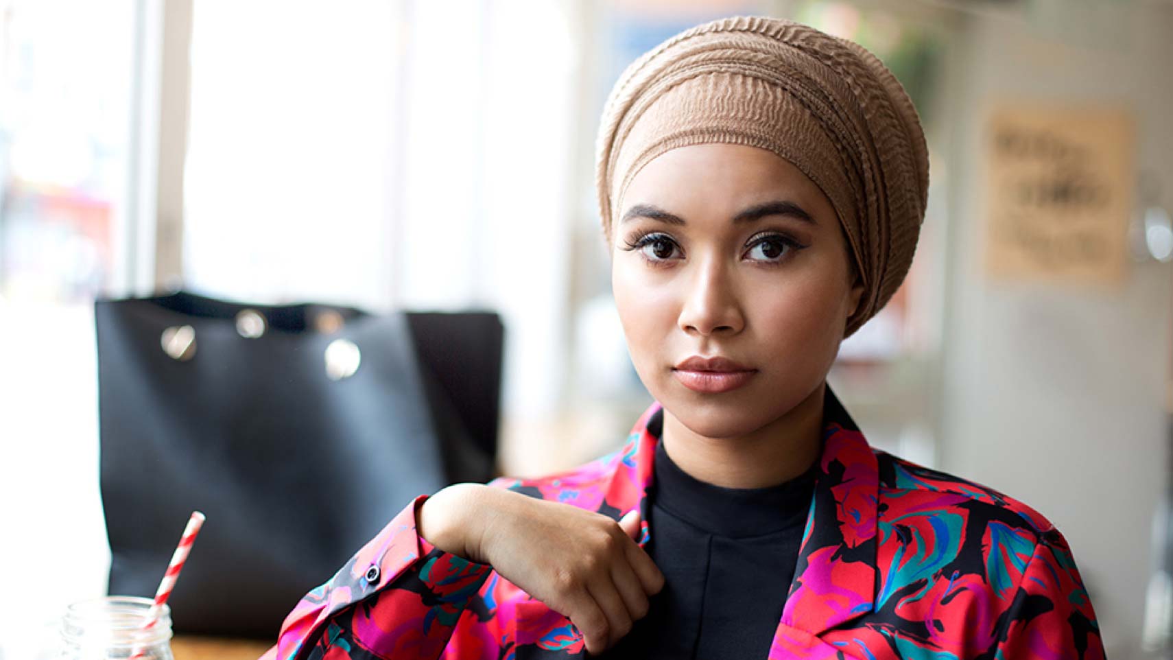 A woman looks into the camera. She is wearing a covering over her hair, a black shirt with a bright, colorful pink jacket over it.