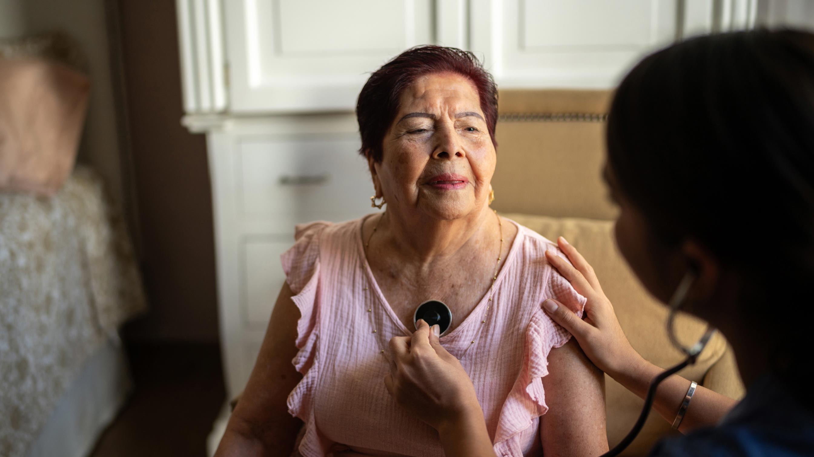 Seated women getting her chest checked with a stethoscope in her home.