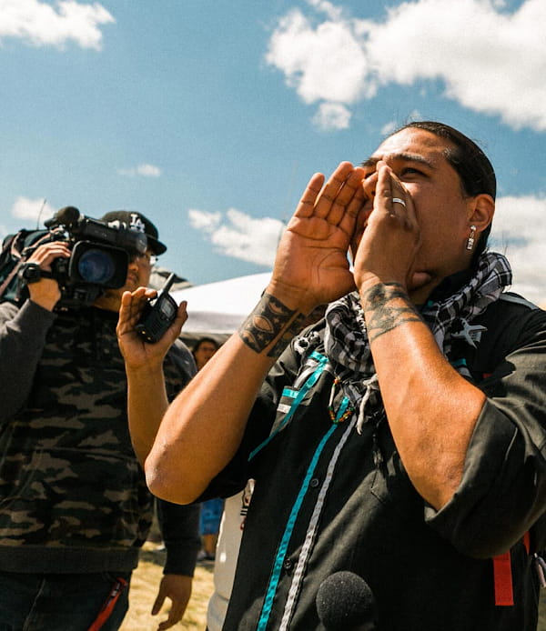 An Indigenous man participating at a NODAPL protest with a camera in the background.