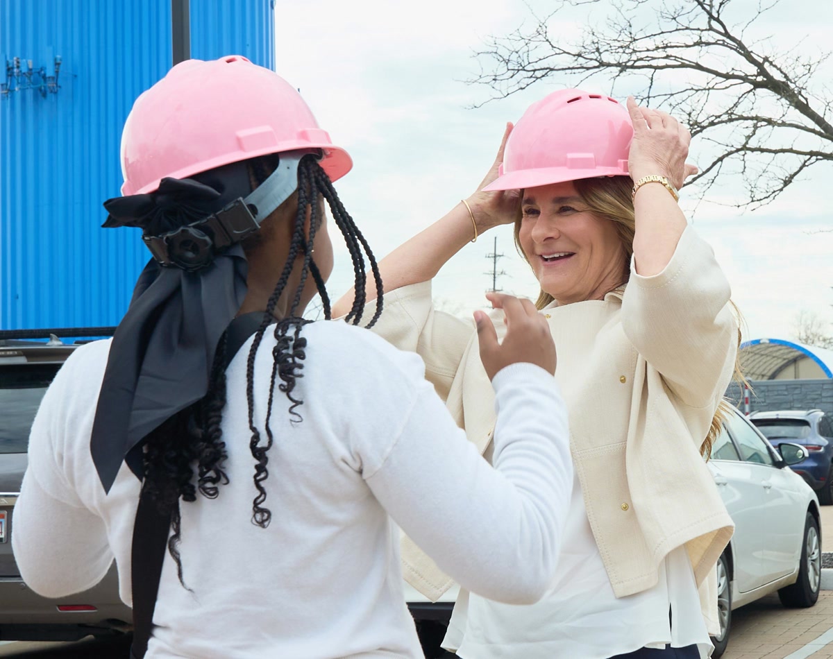 Melinda French Gates puts on a pale pink construction hat with another woman, also in a pink construction hat.