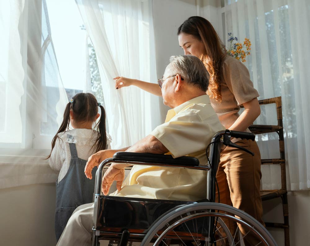 A senior man in a wheelchair looking out of a window with his daughter and young granddaughter.