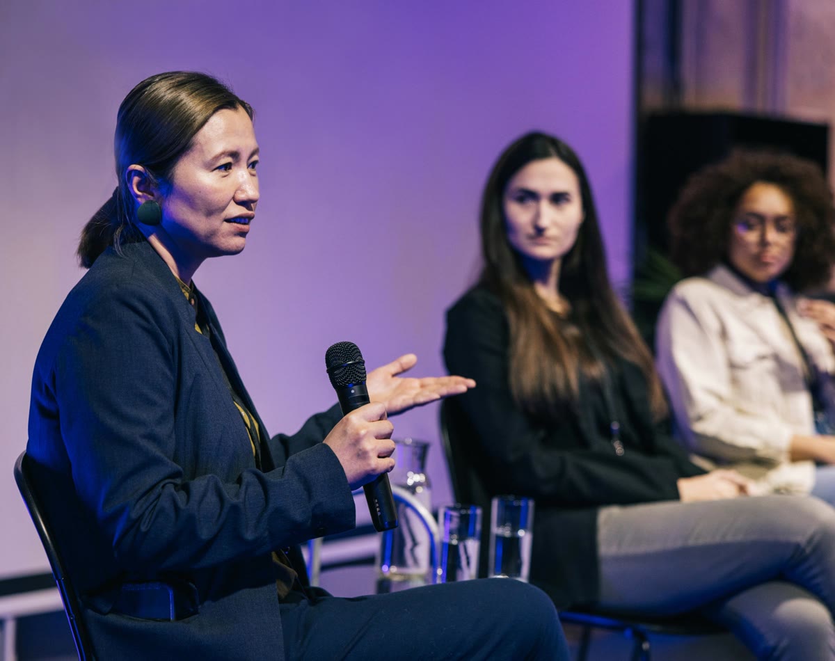 A woman holds a microphone and speaks on stage with two other women at a conference.